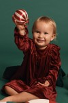 Petite fille souriante tenant boule de Noël en robe Idalia velours bordeaux